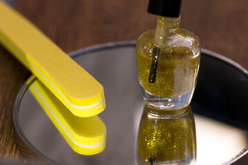 A bottle of translucent nail polish with gold glitter and a yellow nail file on a mirror surface