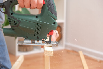 The hands of a master carpenter with an electric jigsaw in his hands cutting off a piece of wood. Male hands using fret saw for cutting wood. A carpenter cuts wood with a jigsaw in a home workshop.