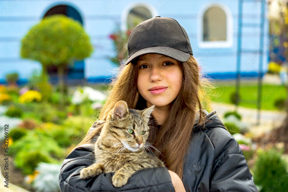 Wall mural Portrait of a girl with a cat in her arms in a black hood on the street.