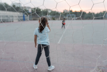 Blurred image. Back view of female child goalkeeper ready to catch a soccer ball stand on soccer field in football goal. Selective focus on girl.