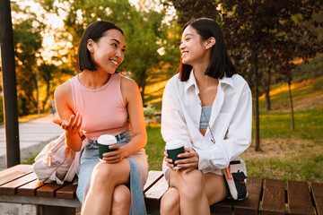 Two asian women talking and drinking coffee while sitting on bench