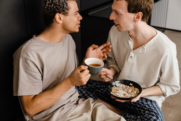 Happy multiracial gay couple talking while having breakfast at home