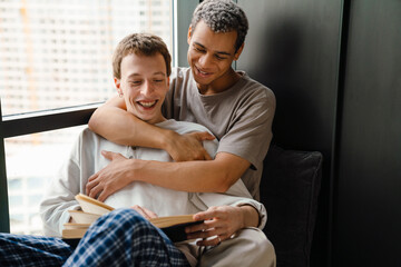 Happy young gay couple hugging and reading book together at home