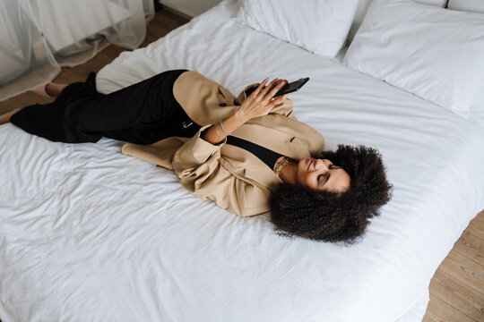Young curly businesswoman using cellphone while lying in bed at home