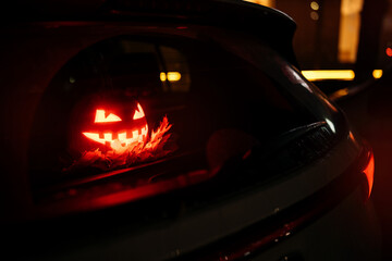 halloween concept. A pumpkin with a glowing red face inside a car at night. Scares passers-by in the parking lot and on the road. - obrazy, fototapety, plakaty