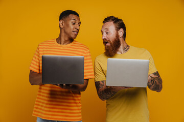 Two multinational men holding laptops and peeking while standing isolated