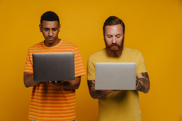 Two multinational men working with laptops while standing isolated
