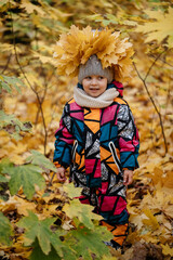 A child in colored overalls and a hat with a wreath of yellow leaves on his head. Walking in the woods in October.