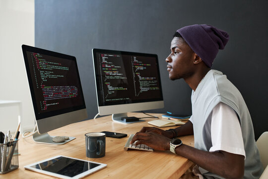 Young serious African American male programmer working over new software in front of computer screens with coded data in office