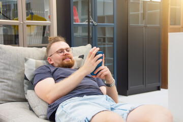 Young caucasian red man with a beard and glasses lies on a sofa holding a smartphone in his hands. Leisure, online shopping, news.