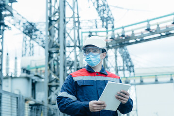 A masked power engineer during a pandemic inspects the modern equipment of an electrical substation...