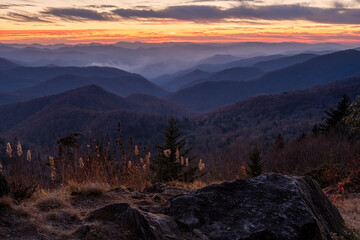 A beautiful and colorful sunset in the Smoky Mountains with an orange sky, blue mountains, and a forest fire in the distance. There are rocks and native plants in the foreground. The sky has clouds. 