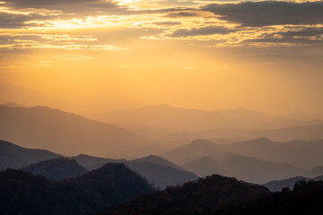 A glowing mountain sunset in the Smoky Mountains taken from the Blue Ridge Parkway. There are clouds in the sky and the hazy atmosphere is glowing orange. There are layers of mountains. 