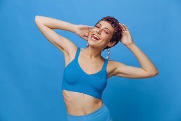 Fototapeta na wymiar Young athletic fashion woman with colored hair and short haircut posing and dancing in blue sportswear smiling and looking at the camera on a blue monochrome background