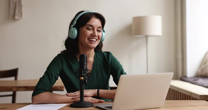 Millennial woman in wireless headphones sit at desk staring at laptop screen, make speech into microphone, record podcast, take part in on-line webinar event, lead online class. Live stream activity
