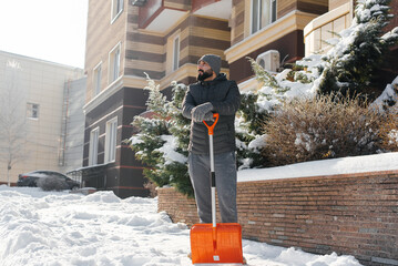 A man stands with a shovel and removes snow in front of the house on a sunny and frosty day....