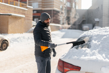 Obraz premium A young man cleans his car after a snowfall on a sunny, frosty day. Cleaning and clearing the car from snow on a winter day. Snowfall, and a severe snowstorm in winter.