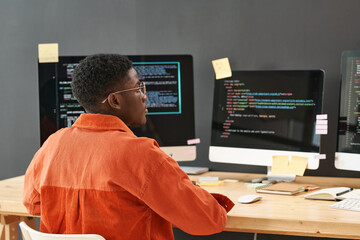 Rear view of young black man concentrating on decoding data on computer screen while sitting by...