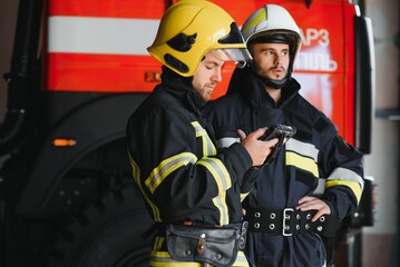 Two professional firefighters with uniforms and protective helmets posing infront of a firetruck.