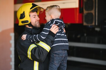 Dirty firefighter in uniform holding little saved boy standing on black background.