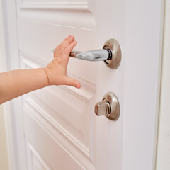Toddler baby opens the door holding the door handle, child hand close-up. White wooden door, metal...