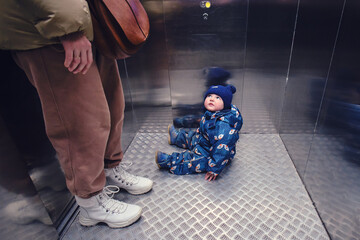 Toddler baby sits on the floor in the elevator next to his mother. The child sat on the floor of the elevator during a trip with a woman mother. Kid aged one year and three months