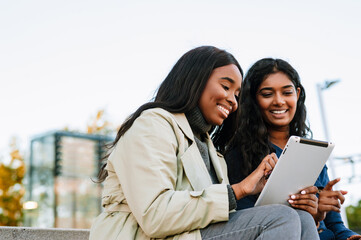 Two young joyful women using tablet computer while sitting at city street