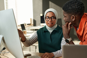 Young confident Muslim woman in hijab pointing at computer screen and looking at African American male colleague during presentation