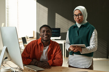 Two young software developers looking at camera while black man sitting by desk and Muslim woman...