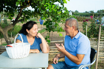 Happy elderly couple sitting at table under green lemon tree and talking while resting after work...