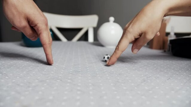 Two people play soccer with their fingers using a small toy ball on the kitchen table.