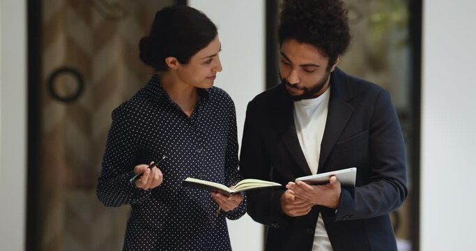 Two Indian male and female office employees walk in modern workspace hallway, discuss common task, using digital tablet device, mentor helps to intern with work during informal meeting during break