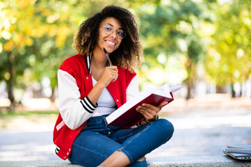 Fototapeta premium Beautiful african female college student reading a book on a bench in a park