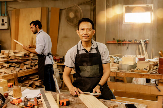 Portrait accomplished senior Asian male carpenter at a furniture factory sits at a desk full of woodwork looking at the camera with a worker standing behind him to select quality wood : Soft focus