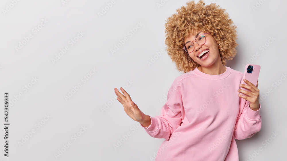 Wall mural Happy young woman with curly hair dances and holds mobile phone expresses authentic positive emotions wears spectacles and casual jumper smiles broadly isolated over grey background blank space - Wall murals