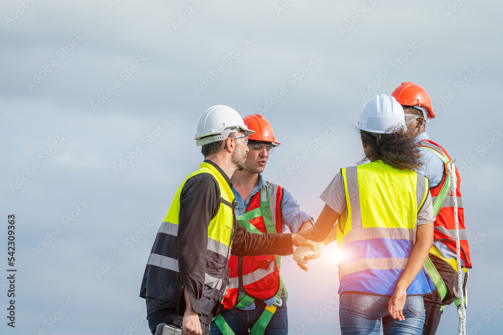 Poster construction workers in protective helmets are shaking hands while working on solar cell on roof fac