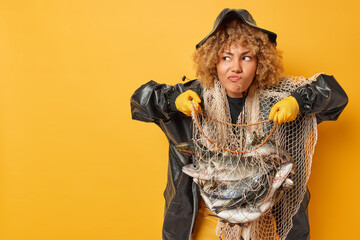 Horizontal shot of serious curly haired female angler holds her trophy goes fishing for hobby frowns face isolated over yellow background empty space for your promotional content. Leisure concept