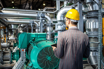 Power plant engineer standing by gas engines inside energy production factory and checking results on digital tablet.