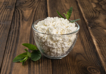 Fresh cottage cheese in a glass bowl and green leaves on a wooden table.