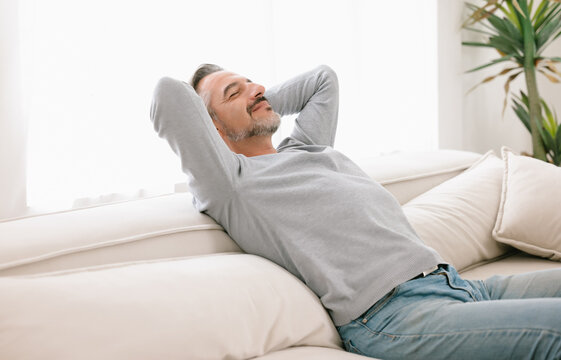 Relaxed man enjoying rest lying on comfortable couch with the hands on the head in living room at home. Calm middle-age man breathing fresh air enjoying no stress free peaceful weekend in house.