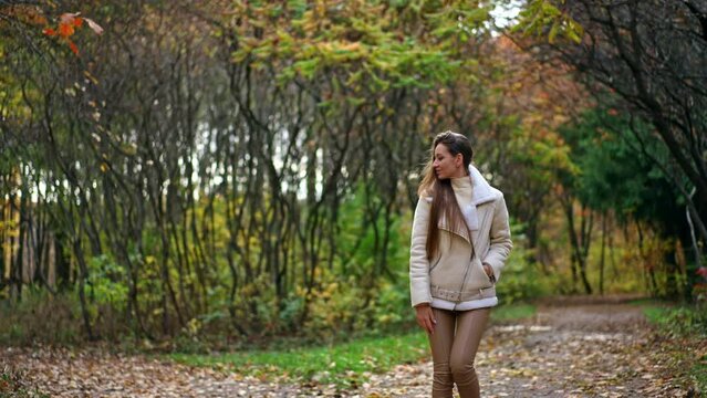 Smiling positive lady in white jacket and tight pants walks backwards looking into camera. Woman turns around and waves her hair and walks away by the road in autumn park.