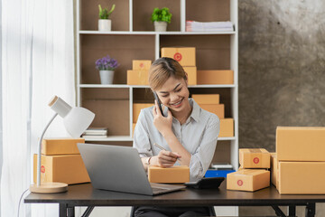 
Smiling Asian woman looking at camera and using laptop to check package delivery information before sending it to customer. Small sme business entrepreneur. Online selling concept.