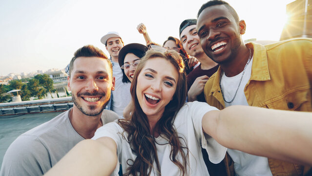 Attractive young woman in sunglasses is taking selfie with friends on rooftop, girl is holding camera and posing while her mates are having fun making funny faces and gestures.