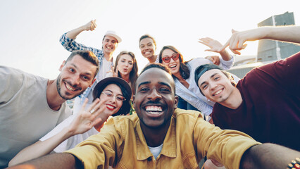 Point of view shot of handsome African American man taking selfie with his happy friends, people are looking at camera, smiling and posing with bottles at funny roof party. - 542325740