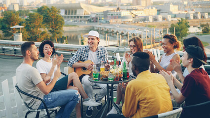 Students are having fun on rooftop playing the guitar, singing and dancing sitting at table on rooftop enjoying free time. Modern lifestyle, joy and musical instruments concept.