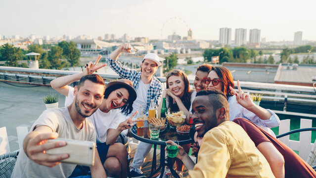 Bearded guy is holding smartphone and taking selfie with joyful friends who are posing with funny faces and hand gestures sitting at table on rooftop having fun.