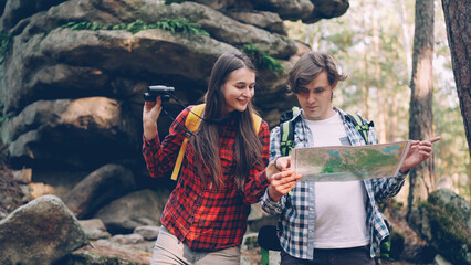 Lost tourists are standing in forest near huge rocks, looking at map then through binoculars and...