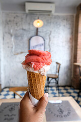 Ice cream cone or gelato held in the left hand. Photographed from the right. With a background of cafe tables and chairs.