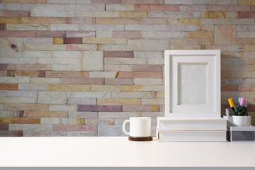 Stylish workplace blank picture frame coffee cup, stationery and books on white table against brick wall.