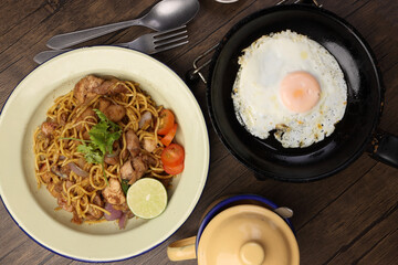 Stair fried yellow noodle traditional mee gorng tomato parsley lime on enamel metal plate cup sunny side up egg in frying pan fork spoon dark soya sauce